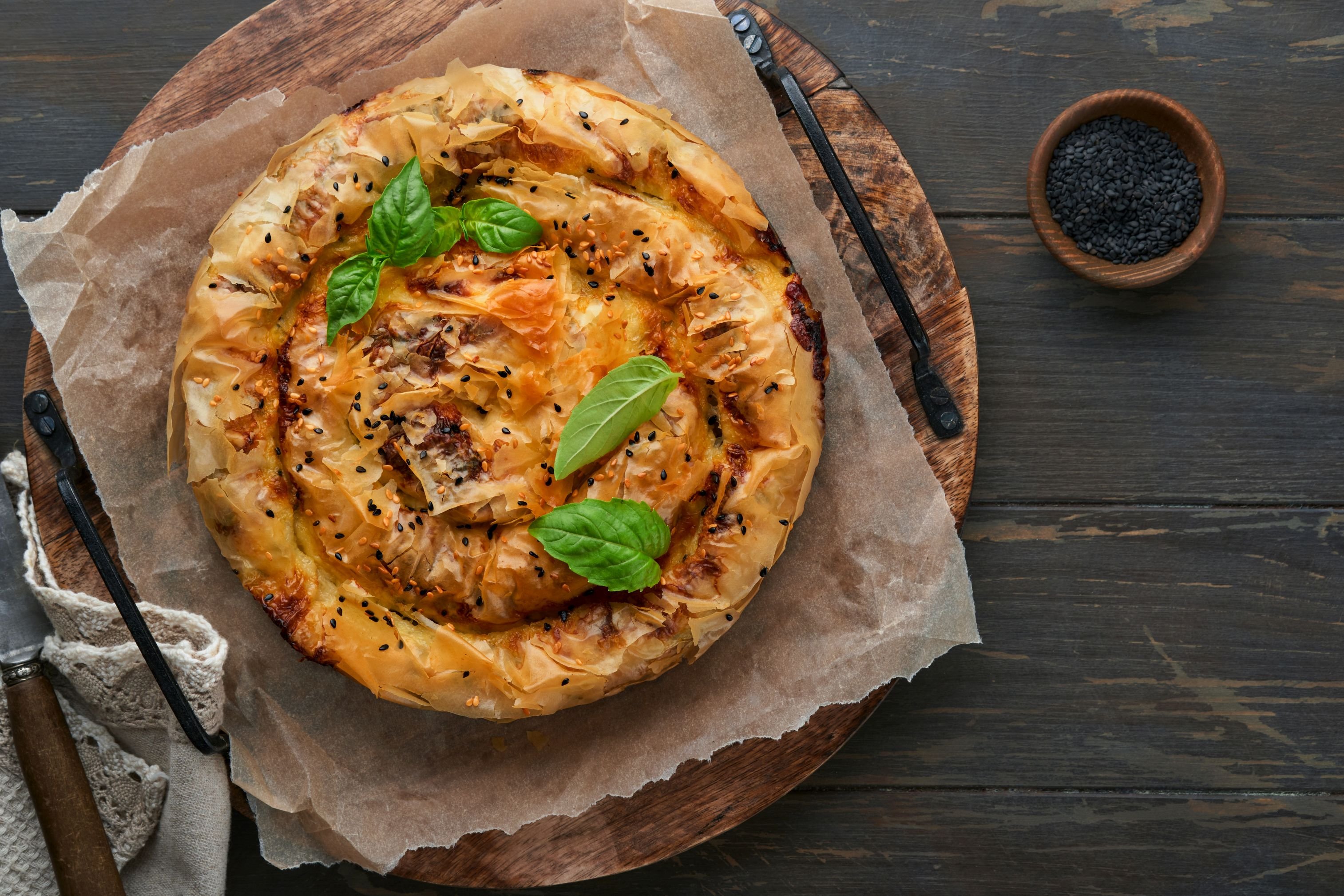 Appetitliches rundes Gebäck, frisch gebacken, aufgegangen, mit Sesam und Basilikum garniert, auf Backpapier, rustikalem Holzbrett und Holztisch