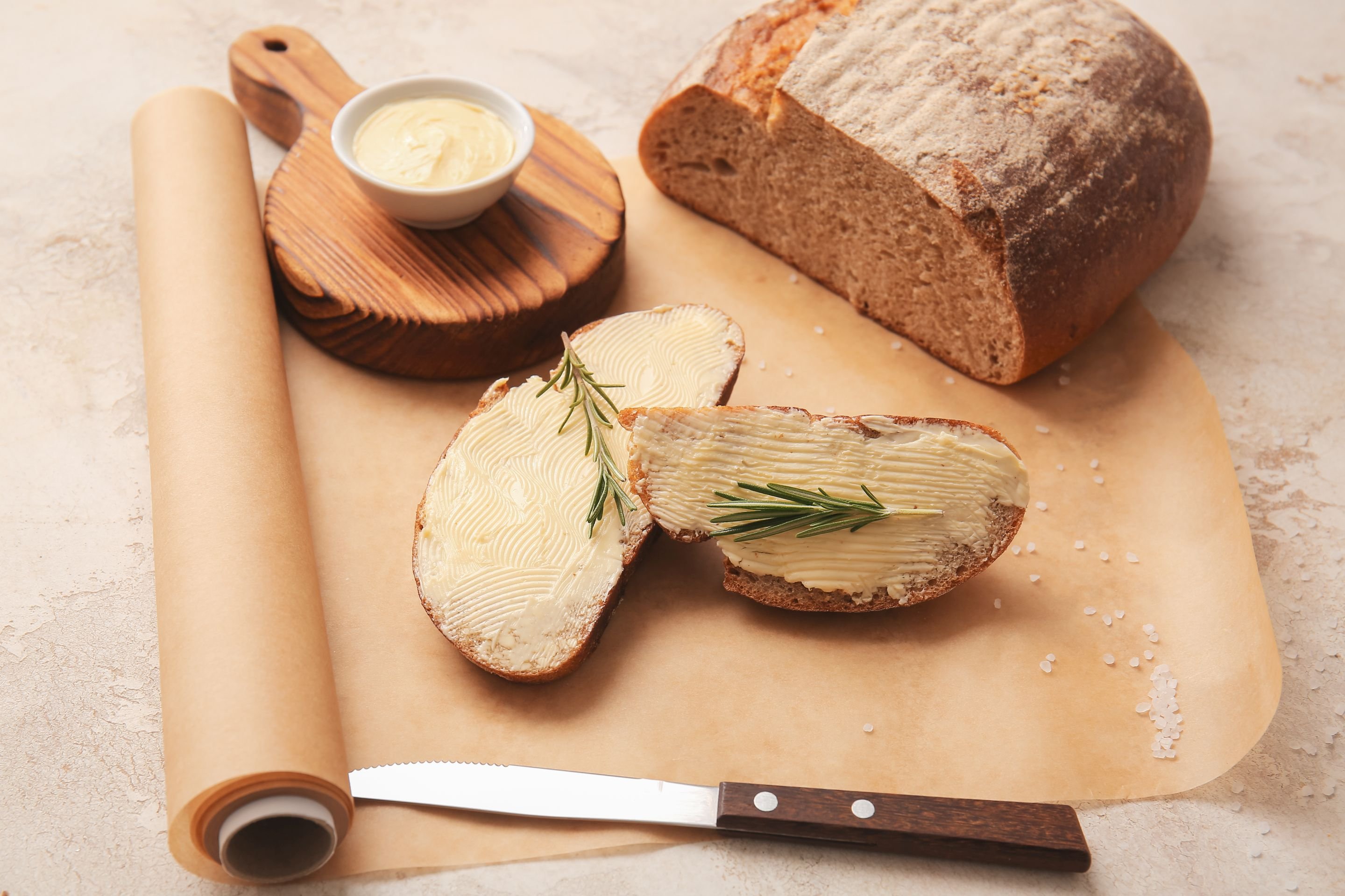 Brotlaib, Scheiben mit Butter, kleines rundes Holzbrett mit Butterschälchen. Darunter Papierrollemit grobkörnidem Salz und Messer