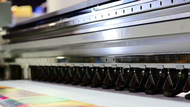 Metallstreben einer Druckmaschine dominieren die rechte Seite, quer, im Vordergund unten links: ein bunt bedruckter Bogen